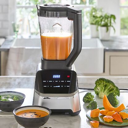 Blender avec soupe chaude au potiron, accompagn&eacute; de deux bols fumants et de l&eacute;gumes coup&eacute;s sur le plan de travail.