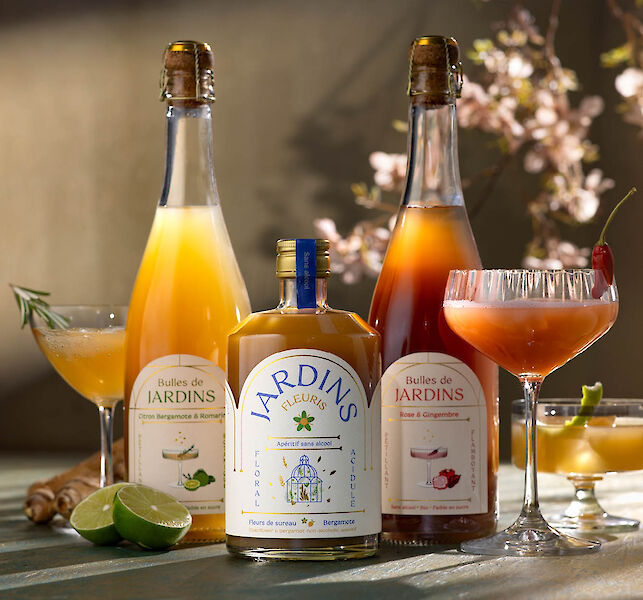 Bouteilles d&rsquo;ap&eacute;ritifs sans alcool &laquo; Jardins &raquo; avec cocktails pr&eacute;par&eacute;s, citrons verts et fleurs d&eacute;coratives autour.