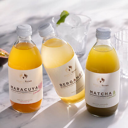 Trois limonades bio en bouteilles : maracuj&aacute;, bergamote et matcha, dispos&eacute;es sur table en marbre avec verres et gla&ccedil;ons.