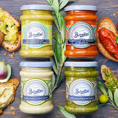Quatre tartinades Bornibus en bocaux (courgette-curry, tomate-basilic, artichaut, olives vertes) avec pain et herbes autour.