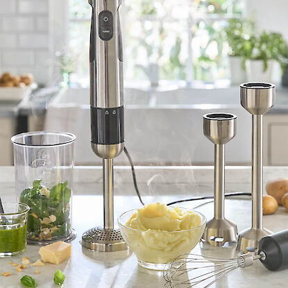 Mixeur plongeant en inox avec accessoires, accompagn&eacute; de pur&eacute;e fumante, ingr&eacute;dients pour pesto et ustensiles sur le plan de travail.