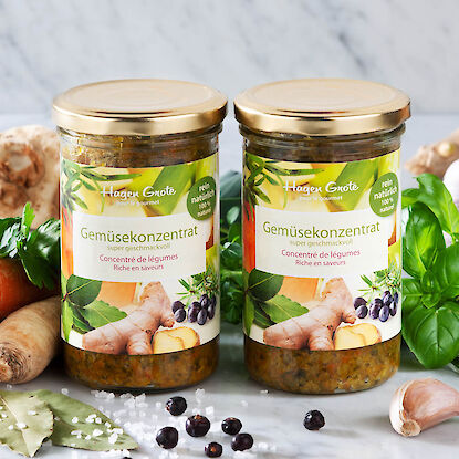 Deux bocaux de concentr&eacute; de l&eacute;gumes Hagen Grote sur marbre, entour&eacute;s de l&eacute;gumes frais, herbes, baies de geni&egrave;vre et sel.
