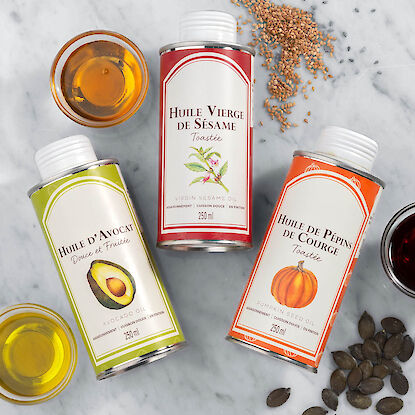 Trois bouteilles d&rsquo;huiles (avocat, s&eacute;same, p&eacute;pins de courge) sur marbre, entour&eacute;es d&rsquo;huiles, graines et p&eacute;pins.