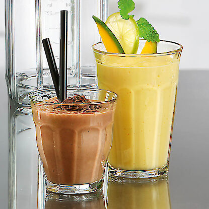 Deux smoothies en verre : chocolat avec pailles et smoothie jaune aux fruits avec citron vert et menthe, devant bol de mixeur.