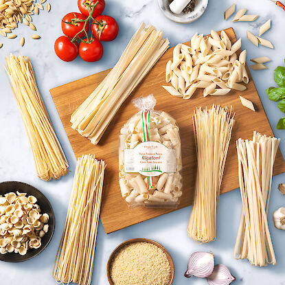 Diff&eacute;rents types de p&acirc;tes autour d&rsquo;un sachet de rigatonis, avec tomates, oignons, riz, herbes et mortier sur surface en marbre.