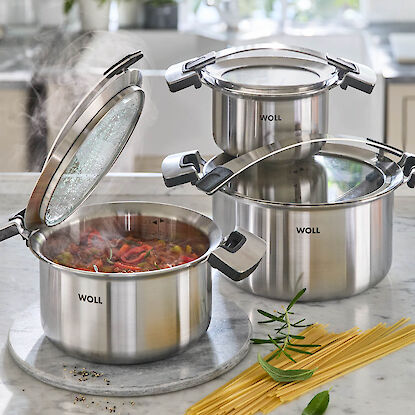 Trois casseroles en inox WOLL, l&rsquo;une avec rago&ucirc;t fumant, accompagn&eacute;es de p&acirc;tes et d&rsquo;herbes sur plan de travail en marbre.
