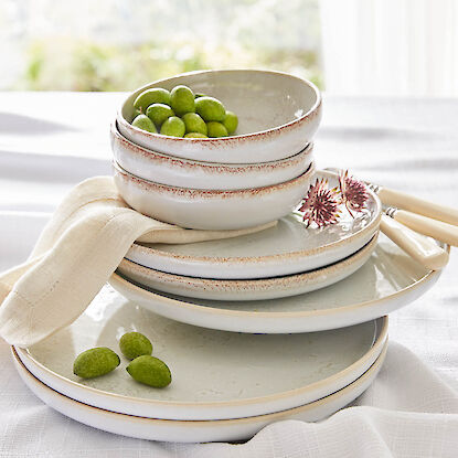 Vaisselle en c&eacute;ramique empil&eacute;e aux tons naturels avec bols d&rsquo;olives vertes, serviette en tissu et couverts sur nappe blanche.