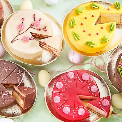 Auswahl festlicher Ostertorten in verschiedenen Farben und Designs, dekoriert mit Blumen, Herzen und Osterdekor, auf Goldplatten.