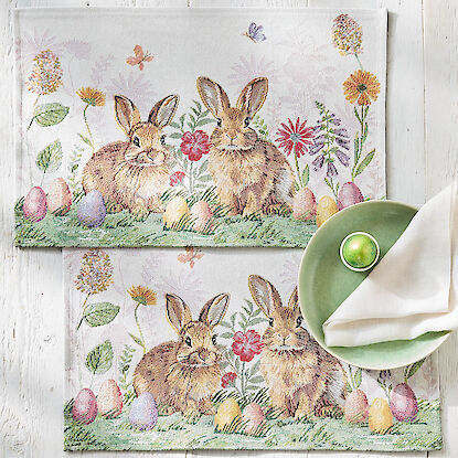 Oster-Tischset mit zwei Hasen zwischen Blumen und bunten Ostereiern, fr&uuml;hlingshaftes Motiv auf hellem Stoff, dekorativ gedeckt.