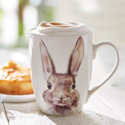 Wei&szlig;e Oster-Tasse mit Hasenmotiv, gef&uuml;llt mit Cappuccino und Milchschaum, auf hellem Holztisch arrangiert.