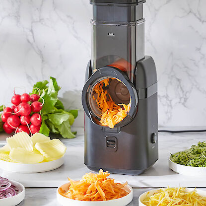 Elektrischer Gem&uuml;seschneider mit rotierender Trommel, raspelt Karotten; umgeben von geschnittenem Gem&uuml;se auf Arbeitsfl&auml;che.