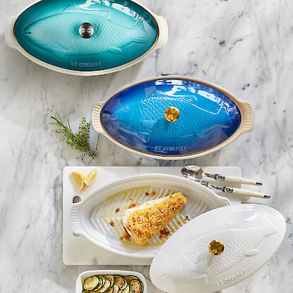 Le-Creuset-Fischterrinen aus Keramik mit Fischrelief, in Blau und T&uuml;rkis, eine ge&ouml;ffnet mit gegartem Fischfilet.