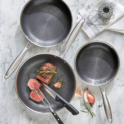 Edelstahl-Bratpfannen mit Wabenstruktur, eine mit gebratenem Steak, Kr&auml;utern und Knoblauch, auf heller Marmorfl&auml;che.