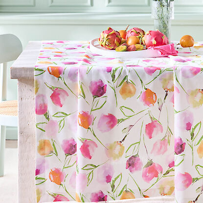 Fr&uuml;hlingshafte Tischdecke mit floralem Aquarellmuster in Rosa und Orange, dekoriert mit Obstschale und Blumen auf Holztisch.