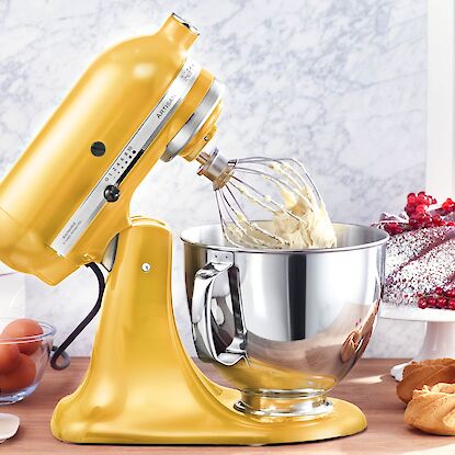 Goldfarbene K&uuml;chenmaschine mit R&uuml;hrsch&uuml;ssel und Schneebesen, beim Aufschlagen von Teig, umgeben von Backzutaten und Geb&auml;ck.