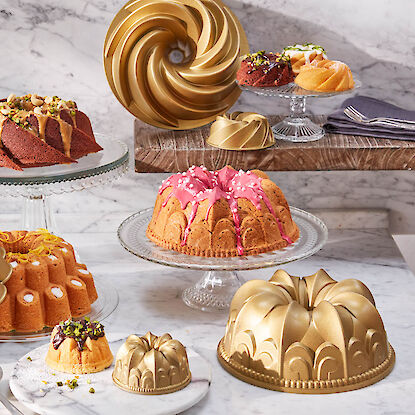 Goldfarbene Gugelhupf-Backformen in verschiedenen Designs, pr&auml;sentiert mit gebackenen Kuchen und Mini-Gugelhupfen auf Marmor.