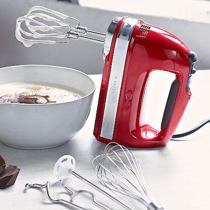 Roter Handmixer mit R&uuml;hrbesen &uuml;ber Sch&uuml;ssel mit Teig, daneben Schneebesen und Knethaken auf heller Arbeitsfl&auml;che.