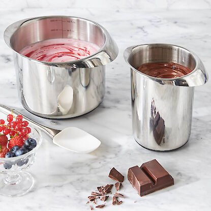 Edelstahl-Schmelzt&ouml;pfe mit Skala, gef&uuml;llt mit Schokolade und Creme, daneben Beeren, Schokoladenst&uuml;cke und Teigschaber.