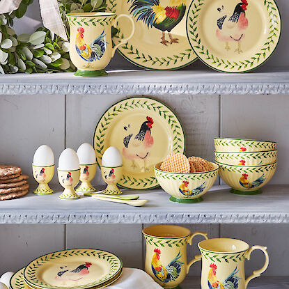 Gelbes Geschirr mit Hahn- und H&uuml;hner-Motiv: Teller, Schalen, Becher und Eierbecher dekorativ arrangiert im Regal.