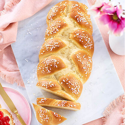 Goldbrauner Hefezopf mit Hagelzucker, angeschnitten auf Marmorplatte, dekoriert mit rosa Tuch und Bl&uuml;ten.