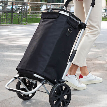 Schwarzer Einkaufstrolley mit gro&szlig;en R&auml;dern, von Person gezogen auf Gehweg im Freien, stabiler Griff und Gestell sichtbar.