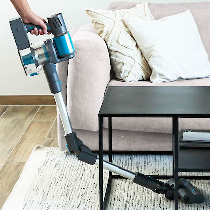 Akkustaubsauger mit Fugend&uuml;se reinigt Sofa und Teppich im Wohnzimmer, Person h&auml;lt das Ger&auml;t seitlich am M&ouml;bel.