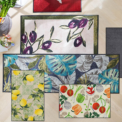 Mehrere K&uuml;chen- und Fu&szlig;matten mit dekorativen Motiven wie Olivenzweigen, Bl&auml;ttern, Zitronen und Gem&uuml;se auf hellem Boden.  Alt-Text Franz&ouml;sisch (&le;125 Zeichen):