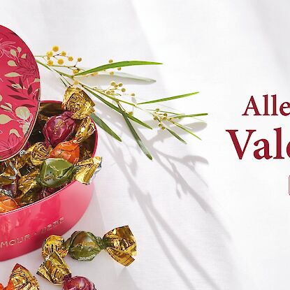Herzf&ouml;rmige Geschenkbox mit Pralinen in Goldfolie, ge&ouml;ffnet auf hellem Hintergrund, mit Aufschrift &bdquo;Alles Liebe zum Valentinstag&ldquo;.