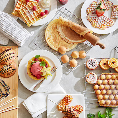 S&uuml;&szlig;er Osterbrunch mit frisch gebackenen Waffeln und Geb&auml;ck, dazu Beeren, Puderzucker, Dessertgl&auml;ser und Waffelrollen auf heller Fl&auml;che.