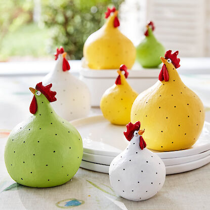 Eine Dekoh&uuml;hnergruppe von 7 H&uuml;hnern in gr&uuml;n, wei&szlig; gelb mit zarten Tupfen aus einem Tisch mit einem Stapel Teller und im Hintergrund einen Blick ins Gr&uuml;ne