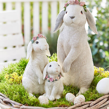 Deko-Hasengruppe bestehend aus 3 Hasen &ndash; gro&szlig;, mittelgro&szlig;, klein &ndash; in einem Osternest mit Moos, gelben Bl&uuml;ten und Vogeleiern in unterschiedlichen Gr&ouml;&szlig;en auf einem Gartentisch mit Garten und wei&szlig;em Lattenzaum im Hintergrund