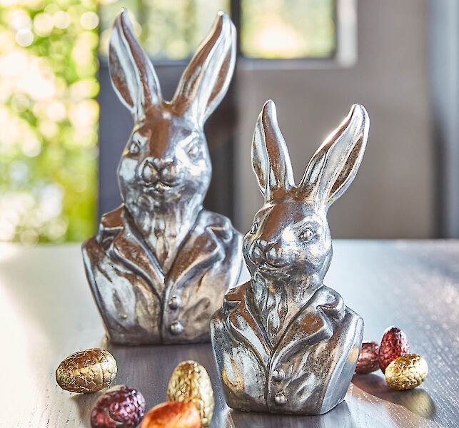 Zwei Silberne Hasenb&uuml;sten auf einem Holztisch mit kleinen Schokoladeneiern in farbigen Alufolien &ndash; gold, Altrosa, orange &ndash; im Hintergrund im unscharfen Bereich ist eine Schiebet&uuml;r in einen sonnigen, gr&uuml;nen Garten