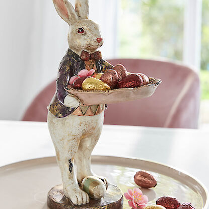 Drei Hasenfiguren mit Blumenkr&auml;nzen in einem Nest aus Moos und Zweigen, dekoriert mit Eiern.
