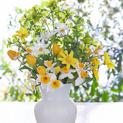 Bunter Fr&uuml;hlingsstrau&szlig; mit verschiedenen Blumen in einer wei&szlig;en Vase auf hellem Tisch vor Fenstern.
