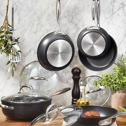 Po&ecirc;les et casseroles suspendues avec couvercles en verre sur plan de travail, accompagn&eacute;es d&rsquo;herbes, d&rsquo;huile et moulin &agrave; poivre