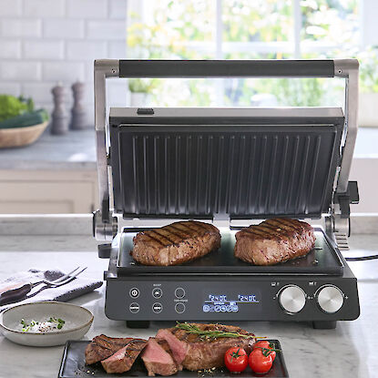 Gril de contact ouvert avec deux steaks en cuisson, steak tranch&eacute; avec tomates pos&eacute; devant le gril