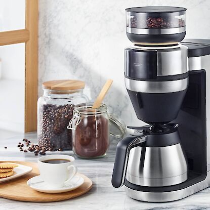 Cafeti&egrave;re filtre avec moulin int&eacute;gr&eacute;, grains de caf&eacute; visibles, carafe et tasse sur plan de travail de cuisine