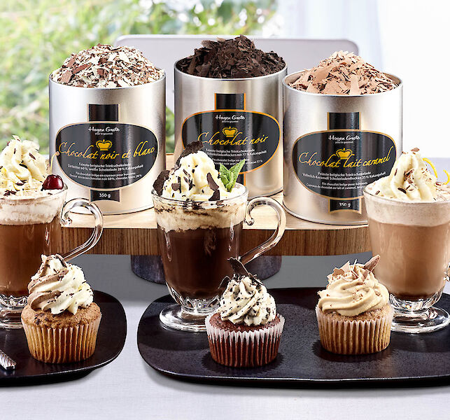 Trois bo&icirc;tes de chocolat chaud en diff&eacute;rentes vari&eacute;t&eacute;s, boissons chocolat&eacute;es avec cr&egrave;me et cupcakes sur table